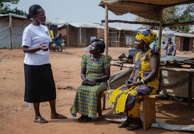 Three women talk to one another in a refugee camp, two sitting down and the other standing.