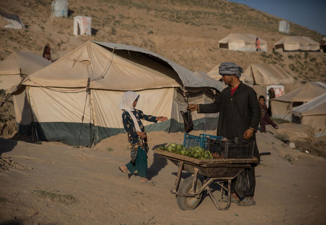 With tents in the background, a man with a fruit cart hands a bag to a young girl.