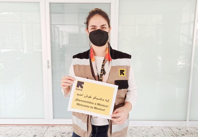Fatima, a home visiting officer for the IRC in Mexico, holds a sign that says "welcome to Mexico" in English, Spanish and Dari. She is wearing a mask and an IRC vest.