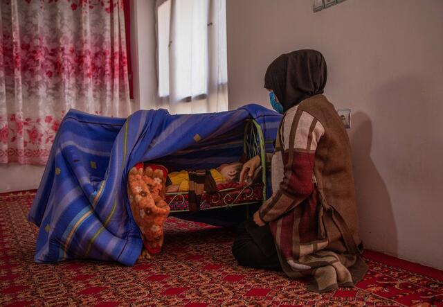 Zulaykha kneels on the ground next to her one-year-old son, who is sleeping bassinet on the ground shielded by blankets.