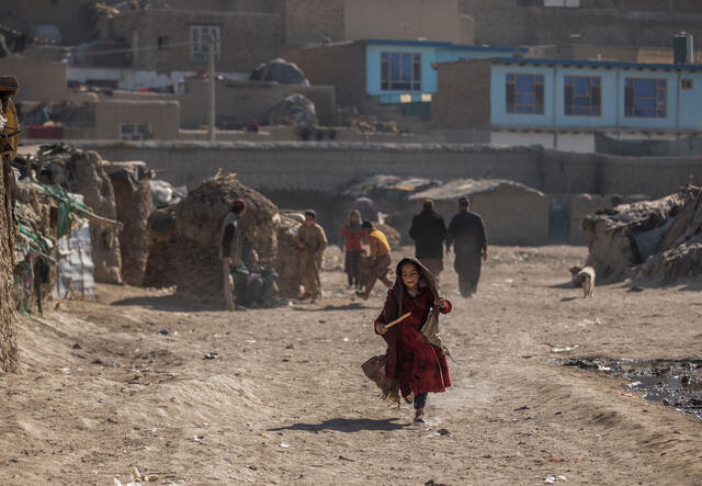 A girl runs through a camp in a desert landscape. There are homes, other children playing and adults walking behind her.