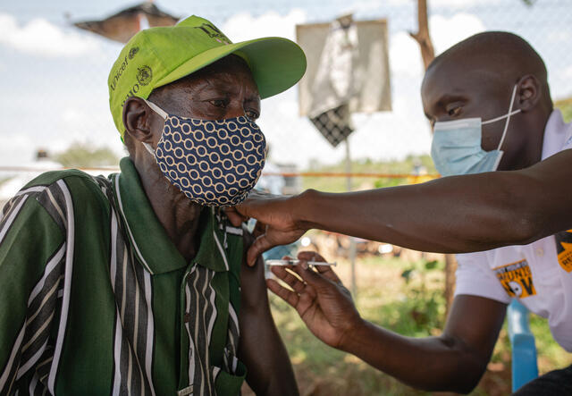 A man wearing a mask and baseball cap gets s COVID-19 vaccine shot from a man also wearing a mask.