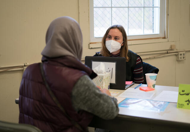 Two woman sit at a table talking to one another with a laptop between them.