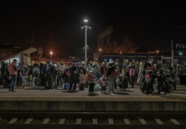 A train platform at night with a large group of people, including many children, waiting. They are wearing winter clothes and many have backpacks.
