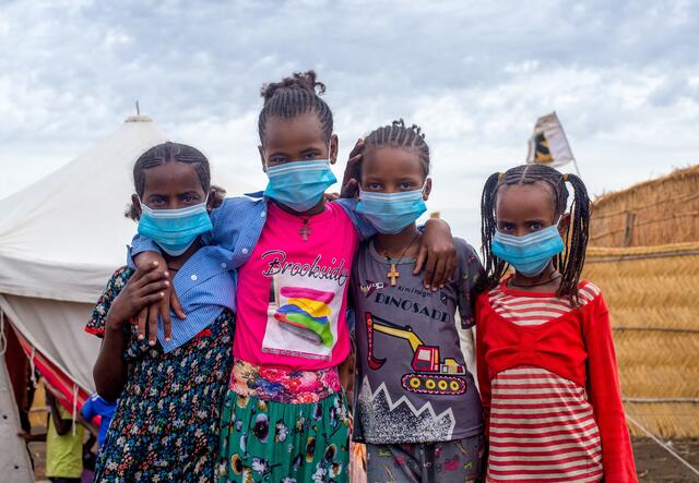 Four children, all wearing masks, put their arms around one another and smile for the camera.