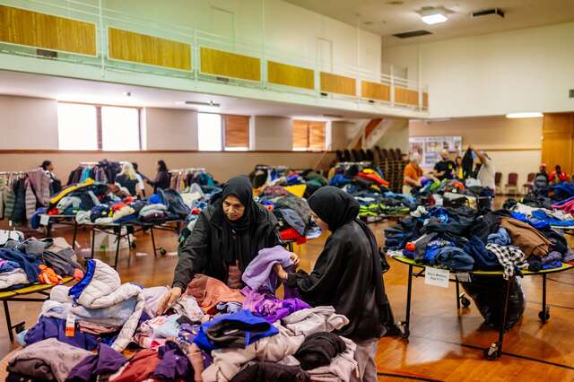 Vaulted ceiling gymnasium filled with tables and clothing racks that are themselves filled with winter coats and jackets; at the front, center table two women wearing hijabs pick through children's winter clothing.