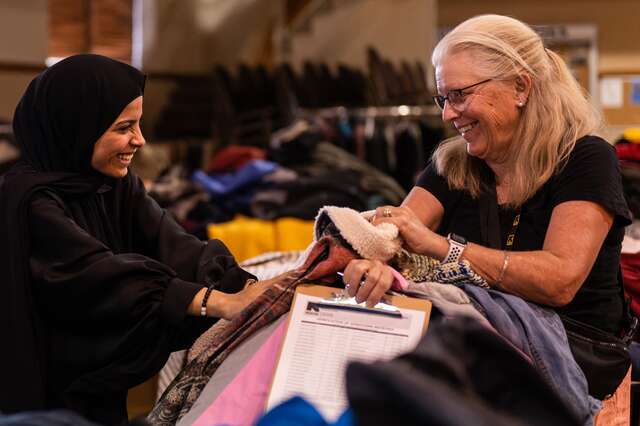 Two women stand facing each other leaning over a pile of winter coats at the IRC's winter clothing drive in 2023.