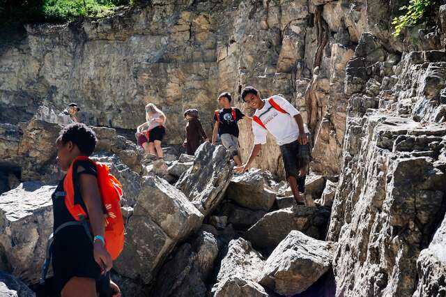 Gray rock face in background and a pile of jagged boulders in the foreground, seven participants in the image in various poses climbing down the rocky mountainside.