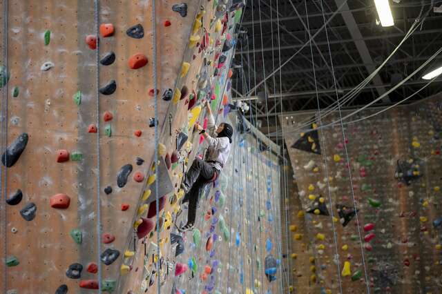 A refugee youth in a hijab in the distance climbs up an indoor climbing wall on her own.