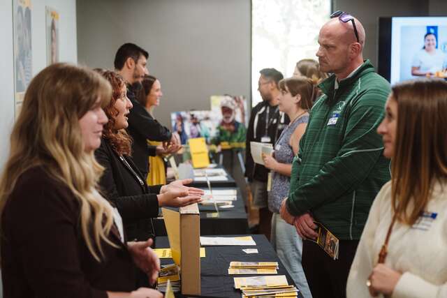 IRC staff members face supporters, answering questions and providing information about IRC programming. IRC staff stand behind tables with black tableclothes (photo left) facing supportings (photo right), including men & women in a variety of spring-time attire.