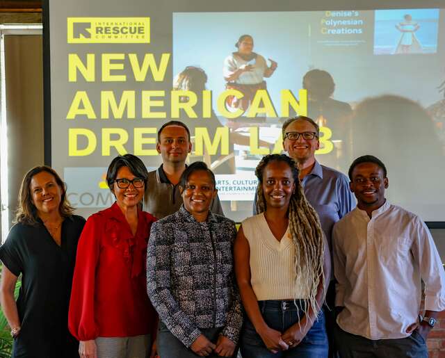 Group photo of New American Dream Lab judges and entrepreneurs standing in front of a projector screen that reads "New American Dream Lab." All are dressed in professional or casual professional attire, looking at the camera and smiling.
