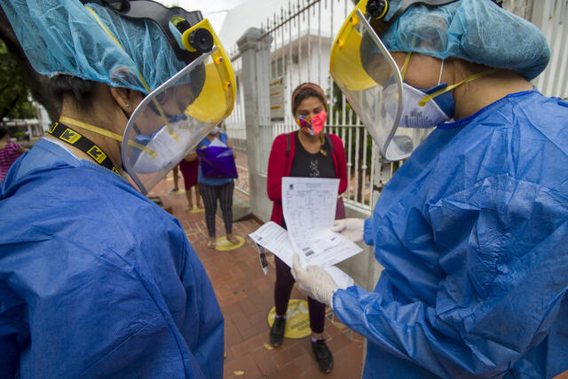 Two IRC health workers in COVID-19 protective gear sign in patients who wait socially distanced on the sidewalk in face masks