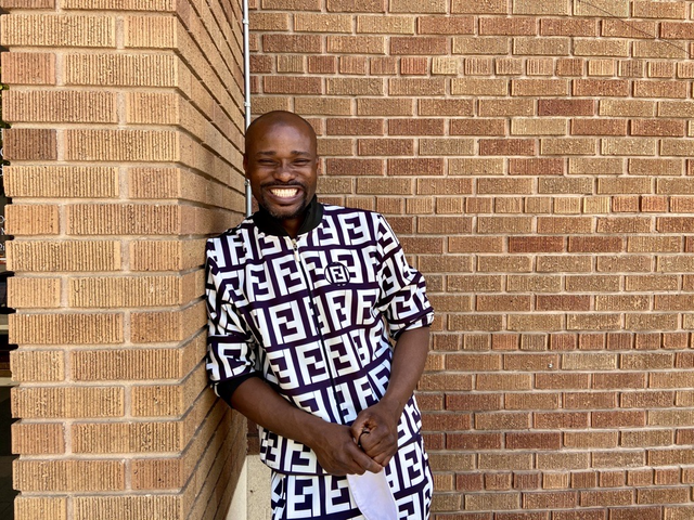 Wilonja casually leans on a brick wall, looking at the camera with a wide grin that lights up his whole face.