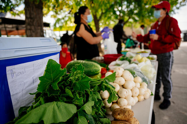 Fresh greens and root vegetables in the foreground on a market table, a customer is being helped by a farmer in the unfocused background