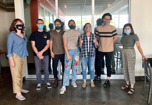Seven staff members of the IRC's Spice Kitchen Incubator, including Emily, stand masked side by side, with smiles and one "thumbs up" pose