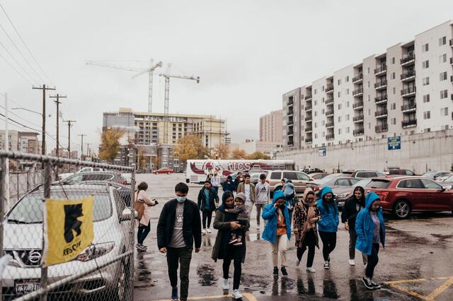 A group of individuals walking through a parking lot with their coats on in the rain through a gate.