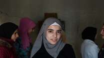 A young woman smiles at the camera in Lebanon. Behind her, other young women speak to an IRC staff member.