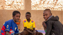 Three boys sit on the floor together in conversation. They look towards the camera.