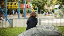 Eine Person sitzt auf einer Bank in einem Park und schaut in Richtung eines Spielplatzes mit bunten Spielgeräten.