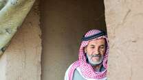 Ein älterer Mann in traditioneller Kleidung steht im Eingang eines einfachen Lehmhauses in Syrien.
