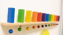 Das Bild zeigt bunte, zylindrische Holzklötze, die auf einem Regal in einer Reihe angeordnet sind, wobei jeder Klotz einer passenden farbigen Vertiefung auf dem Regal zugeordnet ist.