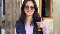 A young woman with long brown hair and sunglasses is looking at the camera and doing a thumbs up with her left hand
