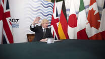Boris Johnson waves his hand as he sits in front of the G7 flags and G7 branding