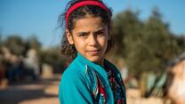 10-year-old Heba looks at the camera. She wears a red head band and green cardigan.