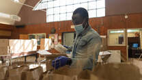 Adam Abakar, a refugee originally from Sudan, works in a food distribution center run by the IRC and World Central Kitchen. He is wearing a mask and gloves, and is putting food into paper bags. 