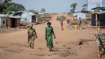 Women's rights activists Foni Grace and Loyce Tabu walk in Bidi Bidi refugeee settlement in Uganda.They are at the forefront of the photo and there are small makeshift homes and a man carrying a bale of hay behind them.