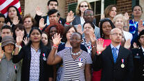 Two new citizens take their oath. They are standing in a large group of people and holding up their right hands.
