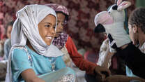 A little girl smiles and looks at a cow puppet while sitting on the ground with other children in Yemen
