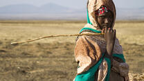 A teenage girl, holding a stick and looking at the camera, stands in a drought affected very dry landscape with mountains in the distance.