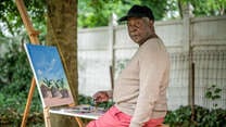 Congolese artist Muyambo Marcel Chishimba site outside at his easel painting a landscape