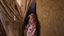 A young girl stands at the entrance to her classroom in Kabul, pulling back a curtain in the doorway and looking into the distance.