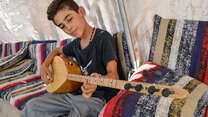 A young boy plays a buzuq in a tent.