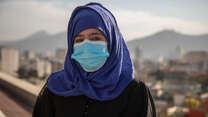 A female Afghan aid worker wearing a surgincal mask stands with a cityscape and distant montains behind her.