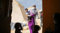 Two children sit on the ground in a tent. Framed in the entrance to the tent is a woman holding a toddler. There are other tents in the background.
