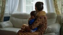 A woman sits on a couch in a living room looking out a window with a toddler on her lap.