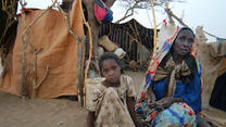 Sudanese refugee woman and girl sitting outside a shelter made of blankets and sheets.