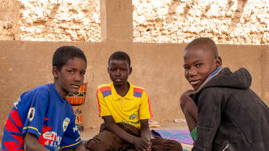 Three boys sit on the floor together in conversation. They look towards the camera.