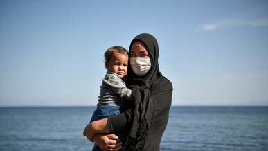 A woman holds a young child in her arms. The two are standing on the beach, in front of the ocean.