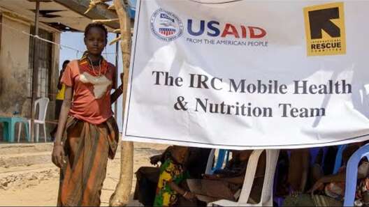 A woman stands next to a sign that says "The IRC Mobile Health and Nutrition Team," with both IRC and USAID logos.
