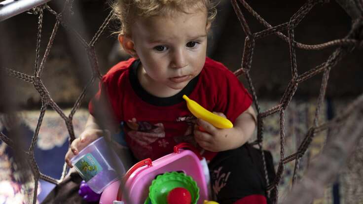 Ein Kleinkind sitzt in einem Netz und hält ein Spielzeug in der Hand, umgeben von bunten Gegenständen, während es neugierig schaut.