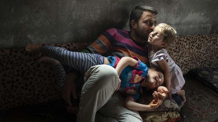 Ein Mann liegt entspannt auf einem Sofa, während zwei kleine Kinder eng an ihn gekuschelt sind; alle drei scheinen friedlich und vertraut miteinander zu sein.