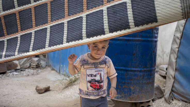 Ein kleines Kind steht in einer provisorischen Unterkunft hinter einer Decke, die als Trennwand dient und hält einen gelben Gegenstand in der Hand.