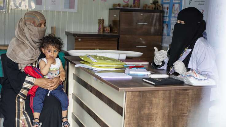 Eine Frau mit Kopftuch hält ein Kind, während sie einer Frau im Niqab an einem Schreibtisch gegenübersitzt.