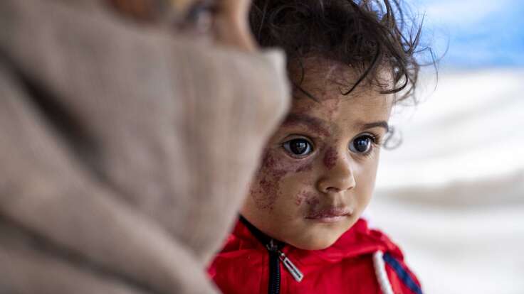 Ein Kind in roter Kleidung schaut nach vorne, während es von einer Person gehalten wird.