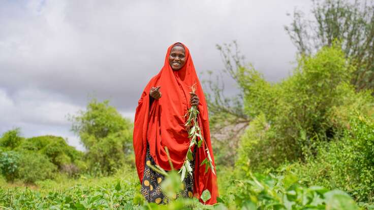 Somalische Frau in rotem Gewand steht lächelnd inmitten eines grünen Feldes.