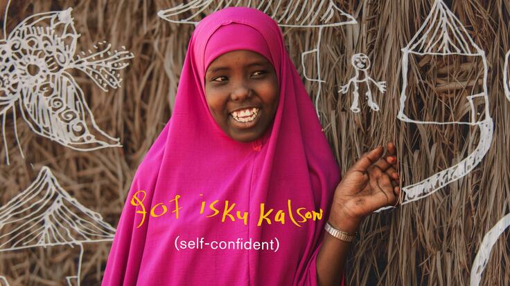 Smiling Asha with her doodle drawings in the background and her handwritten word to describe herself in Somali, translated as “Sociable”